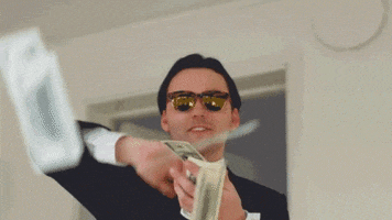TV gif. A cheerful man in a suit with sunglasses makes quick scraping motions over a stack of money, throwing bills into the air.