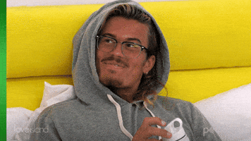 Reality TV gif. Man from Love Island has his hood on and is laying in bed as he laughs.
