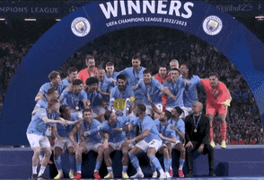 Sports gif. Manchester City football team holds up the 2022/2023 UEFA Champions League trophy under a banner that says "Winners." They all jump up and scream, throwing their arms in the air as fireworks fill the background. 