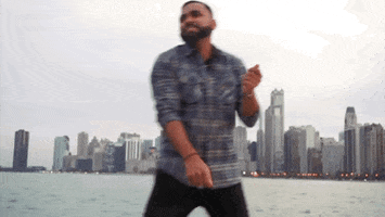 Video gif. Man in a flannel shirt happily vibes and does the dougie in front of a cityscape overlooking the water.