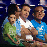 Sports gif. A little boy and three men sit in the audience at a stadium. They suddenly notice they're on the jumbotron and start waving their arms. The boy screams and throws an arm up, then starts dancing with his tongue out, and pulls up his shirt as he moves his hips.