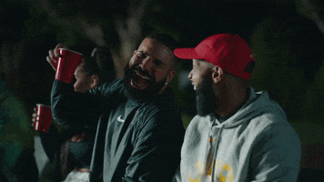 Music video gif. Drake and Odell Beckham Jr. are sitting outside, cackling together. Drake lifts up a red cup and both have their eyes nearly closed and mouths wide open, mid-laugh.