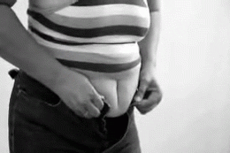 Video gif. A black-and-white scene of a woman's waist as she struggles to button her jeans.