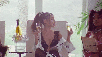 Music video gif. Ariana Grande, Megan Thee Stallion, and Doja Cat are all lounging on beach chairs for their remix 34 + 35. They lift champagne flutes and cheers one another.