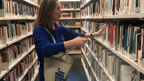 Gif of a woman in a library excitedly grabbing books off the shelf and putting them into a tote bag. 