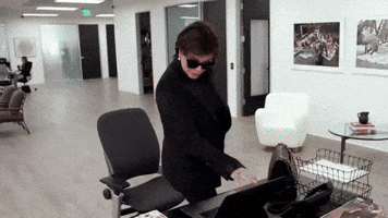Reality TV gif. Kris Jenner on Keeping Up With The Kardashians stands over a desk and closes up a laptop. She has blacked out sunglasses on and she looks suspicious as she goes to grab something on the desk. 