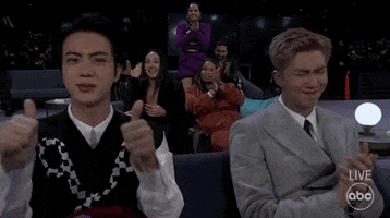 Celebrity gif. Two members of BTS at the 2021 American Music Awards, one giving an emphatic double thumbs-up and the other applauding while shaking his head. 