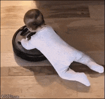 Video gif. A baby in a white onesie clings to the top of a Roomba, sliding along the floor with it as it gently bumps into walls.