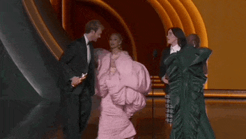 Oscars 2024 gif. Ariana Grande, in an oversized, puffy pink dress, and Cynthia Erivo, in a contrasting forest green dress, run off as Billie Eilish and Finneas O'Connell accept the Best Song award. They awkwardly walk stiffly towards the microphone. 