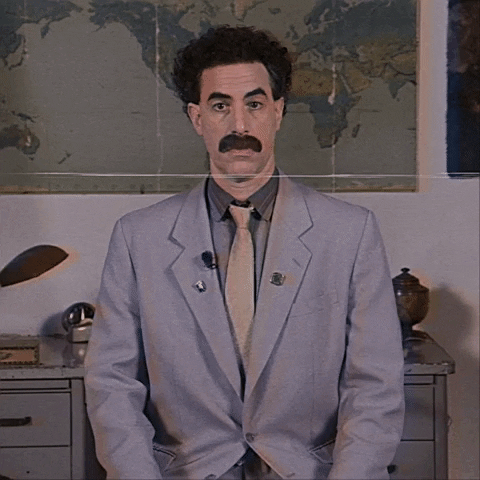 Movie gif. Sacha Baron Cohen as Borat sits in front of a map and raises his two thumbs, waggling his eyebrows and giving an approving nod and toothy grin.