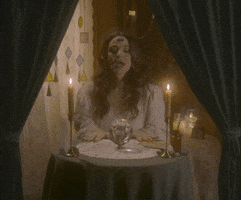 Video gif. Woman dressed as a fortune teller sits at a circular table with candles and tarot cards. She waves her hands over a crystal ball and strikes a dramatic dance pose. Text appears, "I seeee... A happy dance in your future." She looks up and gives us an cheesy, exaggerated grin with an eyebrow raise. 