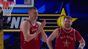 Sports gif. Luka Doncic and Nikola Jokic of the Dallas Mavericks and Denver Nuggets wearing the same red all stars jerseys on the court at the 2024 NBA All-Star Game. Jokic is rubbing his hands together as he smiles and nods at someone offscreen. Doncic has his hands on his hips, smiling and looking amused. 