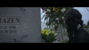 Video gif. Woman slowly approaches a massive tombstone and touches it gently. There are flowers surrounding the tombstone and although it's daytime, it's a very somber atmosphere.