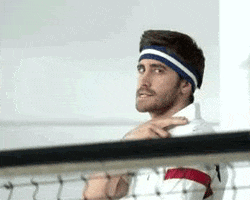 Movie gif. Wearing an athletic headband and a white polo shirt, partially obscured by a tennis net in the foreground, Jake Gyllenhaal glares at us and makes a threatening gesture by sliding two fingers across his neck.