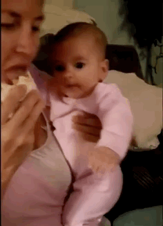 Video gif. Mom holding a baby takes a bite of a burrito, and then the baby leans in with a wide open mouth like she's trying to take a bite.