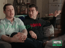 Sponsored GIF. Noah Reid and Dan Levy sit in a living room on a couch. While in discussion, Noah tries to neutralize their situation by turning to Dan to guide the scenario by saying “we’ll be happy with whatever you have planned....RIght David?” Dan, clearly struggling, tries his best and nods but ultimately dismisses Noah’s cue and responds,” Yes….just depends on what’s she’s planned.”