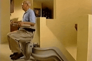 Video gif. An elderly man sits in a motorized stair lift chair as it rounds a corner about to descend a flight of stairs.
