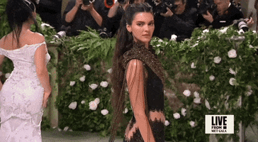 Met Gala 2024 gif. Kendall Jenner wearing a plunging black and bronze vintage Givenchy 1999 Haute Couture gown, posing for the cameras, tries to settle into the right pose.