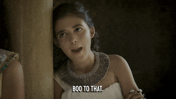 TV gif. Olivia Trujillo as Arsinoe in Drunk History crosses her arms as she leans against a pillar and stretches her neck out to say, "Boo to that," before rolling her eyes.
