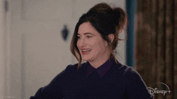 Marvel gif. Kathryn Hahn as Agatha Harkness in WandaVision looks down awkwardly before breaking the fourth wall and staring straight at us with wide eyes. The Disney Plus logo rests in the bottom right corner.