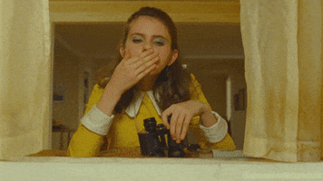 Movie gif. Kara Hayward as Suzy in Moonrise Kingdom next to a window, holding binoculars and glancing down at us with seriousness as she blows a kiss.
