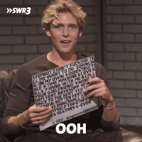 Celebrity gif. Christopher Nissen grins seductively as he fans himself with a sign. Text, "Ooh, it's kinda hot in here." 