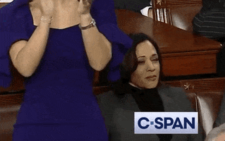 Political gif. While a person in a blue dress next to her applauds, Kamala Harris looks on without emoting.