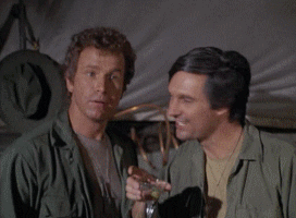 TV gif. Alan Alda as Capt. Benjamin and Wayne Rogers as Capt. John in MASH. They both hold up cocktail glasses and point at someone before moving towards them to cheers together.