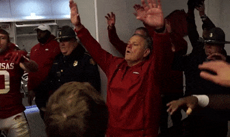 Sports gif. Sam Pittman, head coach for the Arkansas Razorbacks, raises his hands in the air and waves his fingers before doing a fist pump. Everyone follows his lead in the team cheer.