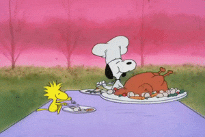 Cartoon gif. Characters Snoopy and Woodstock tuck into a Thanksgiving meal on a blanket spread out on the ground.