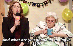 SNL gif. Vanessa Bayer as a news reporter, holds a microphone and smiles. She sits in a room decorated with balloons and a sign that says "Happy Birthday." She turns to a man dressed as an old woman in a wheelchair and asks, "And what are you gonna do to ring in 110?"
