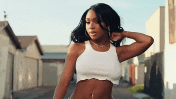 Music video gif. In a wide alley on a sunny day, wearing a white half-shirt and hoop earrings, Normani gives us a friendly stare and does a hair flip.