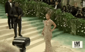 Met Gala 2024 gif. We zoom in on Jennifer Lopez posing with one hand on her hip and a stoic yet confident expression. She's wearing a nearly-see-through Schiaparelli sleeveless gown with a plunging v-neckline that culminates in a pattern that flares out from her shoulders like butterfly wings. She's wearing a prominent diamond necklace, also resembling feathers. 