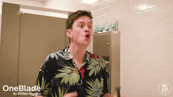 Video gif. A young man hypes himself up in front of a public bathroom mirror, holding a men's electric razor.