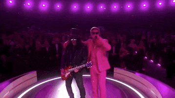Oscars 2024 GIF. Ryan Gosling, dressed in a Barbie pink tuxedo and sunglasses, his arm around Slash, as we circle them performing I’m Just Ken.