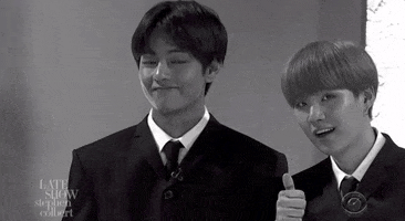 TV gif. From the Late Show with Stephen Colbert, black and white image of V from BTS smiling and cradling his hands around his face while Suga leans in giving a thumbs up.