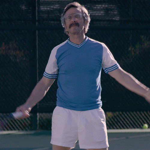 TV gif. Marc Maron as Sam Sylvia from GLOW is dressed appropriately as he stands on a tennis court holding a racquet and shrugs with disappointment.