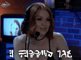 Video gif. Attractive redhead woman wears a gamer headset and smiles sweetly, saying, "I missed you," which appears in text in multiple languages. The background has ambient blue lighting and a retro music video plays on a CRT television.