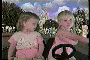 Video gif. Two small blonde girls appear to be driving a small car down a street at Disney World, and the passenger pushes the driver out of the car and takes the wheel.