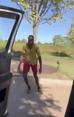 Video gif. We are sitting in a car with the car door open. A man jumps, almost like a frog, sideways alongside the car. He smiles at us and paces his hands on his thighs as he hops along.