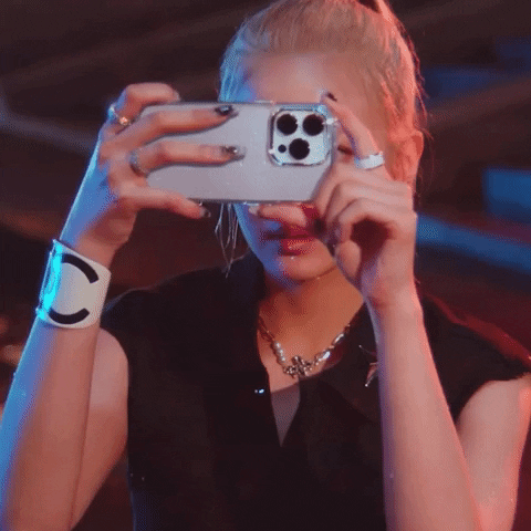 Celebrity gif. Lia from Itzy holds up her phone and takes a picture before putting her phone down and grinning.