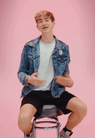 Video gif. Cash Baker, an influencer, is sitting on a stool explaining something and gesturing with his hands.