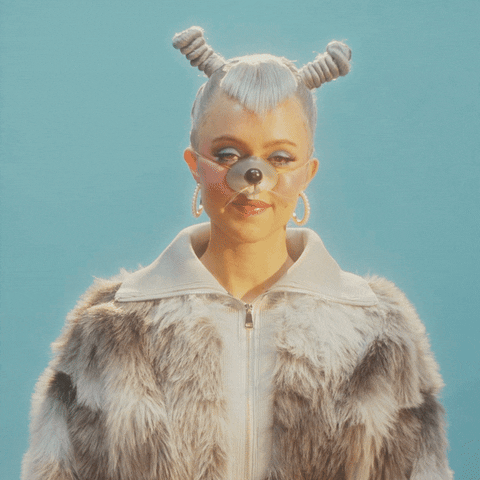 Video gif. Young woman wears a furry jacket and dog nose with platinum hair styled in tight coils and funky bangs against a teal background. She waves at us with both hands, smiling as she says, "Have a great day!'