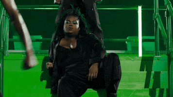 Video gif. Confident and cool, a model on the Savage X Fenty show sits on stage amongst dancers.