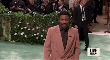 Met Gala 2024 gif. Closeup of Donald Glover smiles and waves then tugs on the lapel of his tan suit, paired with a dark-brown shirt and matching tie.