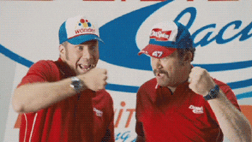 Movie gif. Will Ferrell as Ricky Bobby and John C Reilly as Cal Jr. in Talladega Nights look at us and fist bump as they yell, “Shake and Bake!”