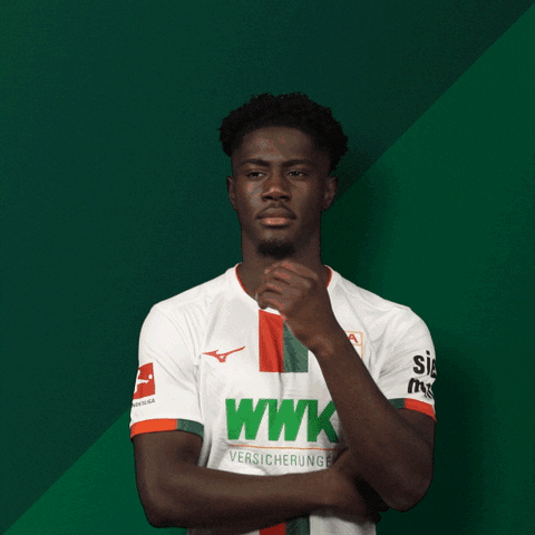 Sports gif. Augsburg soccer player Nathanal Mbuku wears his team's jersey as he leans against a green wall, looking off and pensively stroking his chin in quiet reflection.