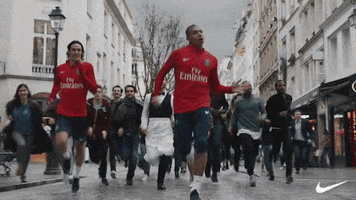 Ad gif. Two men wearing red soccer jerseys run down a city street, leading a crowd of people running with them and inviting more onlookers who join.