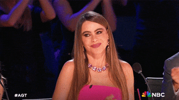 Reality TV gif. Sofia Vergara seated in a judges chair on "America's Got Talent," raising her hands over her head with a big friendly cheer as the audience claps behind her.