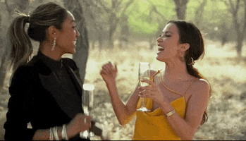 Reality TV gif. Tayshia Adams and Kaitlyn Bristowe from the Bachelorette stand next to each other a sunny forest clearing. They laugh as they toast their white wine, as if to say "cheers!"